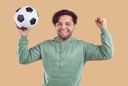 Téléchargez les photos : Joueur de football joyeux s'amuser pendant l'entraînement. Portrait de jeune homme heureux en sweat à capuche vert menthe isolé sur fond de couleur beige tenant ballon de football, souriant, acclamant et levant le poing - en image libre de droit
