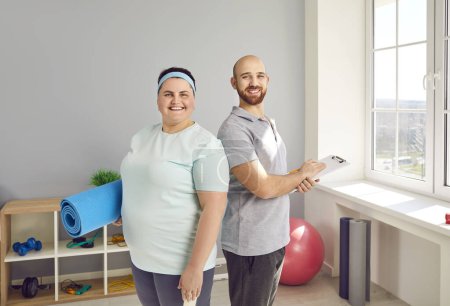 Happy overweight woman together with personal fitness trainer. Portrait of joyful motivated young fat girl with workout mat and cheerful fit man with clipboard standing in gym with sports equipment