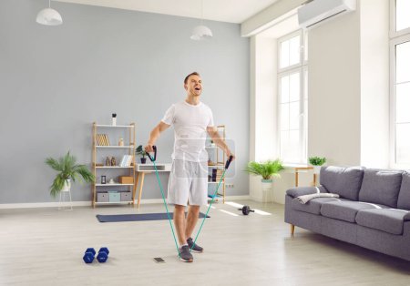 Photo for Young purposeful man at home doing sports strength training using rubber resistance band. Man in t-shirt and shorts shouting while stretching elastic band in living room. Home fitness training concept - Royalty Free Image