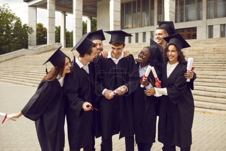 Group of a smiling happy multiracial international graduates students hugging and having fun in a university graduate gown holding diploma outdoor. Education and graduation concept.