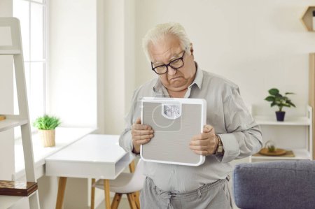 Photo for Upset retired grey-haired senior man holding weighing machine standing in the living room at home. Elderly male frustrated person looking sadly on his scales. Weight loss and diet concept. - Royalty Free Image