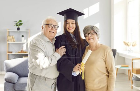 Photo for Young happy girl with diploma in hands in black university graduate gown standing with her senior old parents proud of her in living room at home looking at camera. Graduation and education concept. - Royalty Free Image