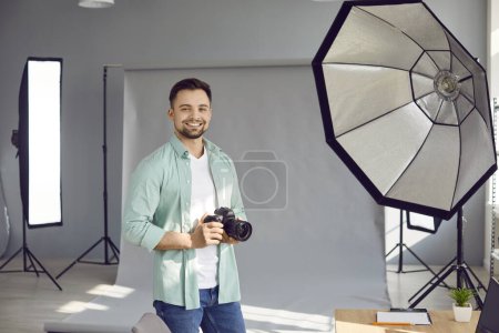 Foto de Retrato de feliz fotógrafo masculino. Joven alegre y sonriente con camisa casual sosteniendo cámara fotográfica y de pie en el estudio con fondo gris y equipo de iluminación. Concepto de fotografía profesional - Imagen libre de derechos