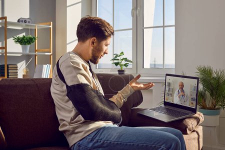 Man with physical injury consulting online doctor on telemedicine clinic website. Young man with broken arm sitting on sofa with laptop computer and talking to doctor via video call