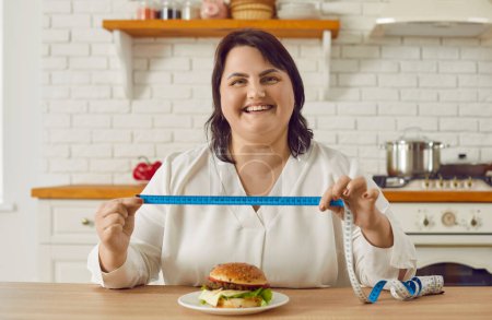 Téléchargez les photos : Portrait d'une jeune femme heureuse et souriante en surpoids, assise sur la cuisine, tenant un ruban à mesurer avec un hamburger sur la table. Restrictions alimentaires, saine alimentation, amaigrissement et concept de perte de poids. - en image libre de droit