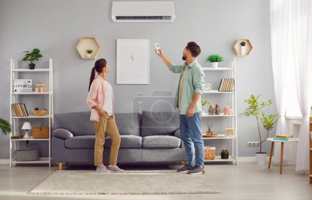 Paar zu Hause genießt den Komfort der Klimaanlage mit modernen Wand-Klimaanlage. Mädchen beobachtet, wie Mann mit Fernbedienung Klimaanlage anpasst und Komfort und Kühle des eigenen Hauses genießt
