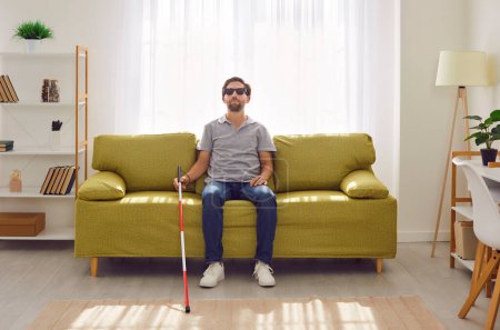 Portrait of a young blind man with stick cane sitting on sofa in the living room at home. Male person in dark glasses with vision problems resting on the couch. Blindness and disability concept.