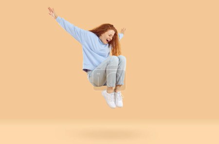 Photo for Happy excited teenage girl is happy jumping high and levitating on beige background in studio. Redhead curly girl in casual clothes having fun jumping with closed eyes and outstretched arms. - Royalty Free Image