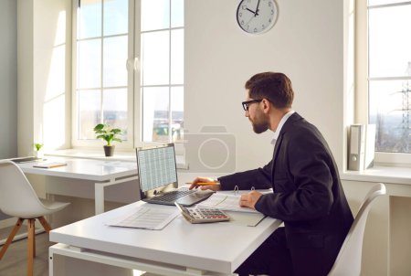 Photo for Business accountant working on a laptop. Man in a suit sitting at his office desk, doing paperwork, using a modern notebook computer, working with digital data sheets and checking some figures - Royalty Free Image