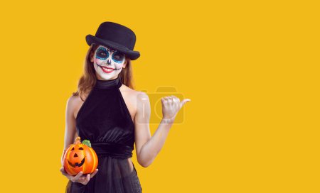 Téléchargez les photos : Jeune fille en costume de carnaval noir avec du maquillage de crâne de sucre debout dans le studio sur fond jaune avec un index pointant la citrouille sur le côté pour copier l'espace. Halloween et concept d'horreur. - en image libre de droit