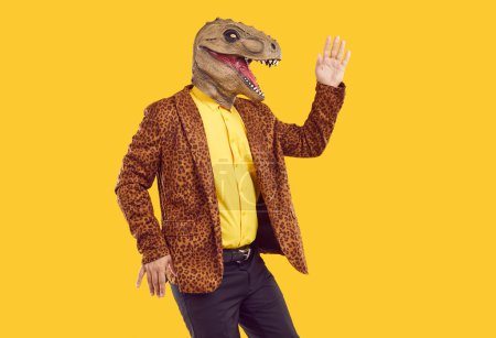 Exzentrischer, lustiger Showman in Gummi-Dinosaurier-Maske tanzt und winkt mit der Hand auf gelbem Hintergrund. Er trug eine Leopardenjacke und ein gelbes T-Shirt. Werbebanner mit flippigem Mann. Party, Maskerade.