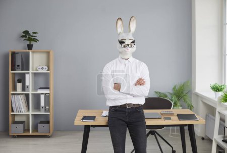 Businessman in rabbit head mask standing with folded hands in office. Funny male office employee, clerk in formal wear, rubber mask and glasses sitting on desk and looking at camera