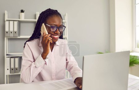 Téléchargez les photos : Portrait de femme d'affaires afro-américaine souriante heureuse comptable assis sur le bureau de son lieu de travail dans un bureau moderne en vêtements décontractés travaillant sur un ordinateur portable et parlant sur un téléphone mobile. - en image libre de droit