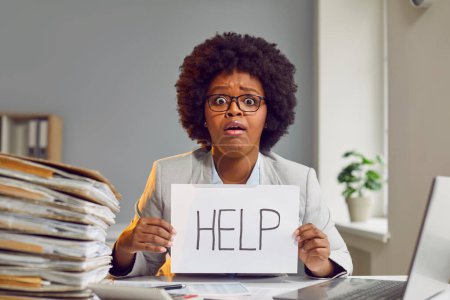 Photo for Very tired Afro American woman in glasses sitting at desk with huge load of paperwork, holding paper sheet with word HELP and looking at camera with funny, sad face expression. Work in office concept - Royalty Free Image