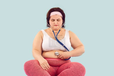 Photo for Large obese plump overweight woman in sports bra and leggings holds stethoscope on her big fat belly, listening to bowel sounds caused by overeating, stomach problems and unhealthy digestive system - Royalty Free Image