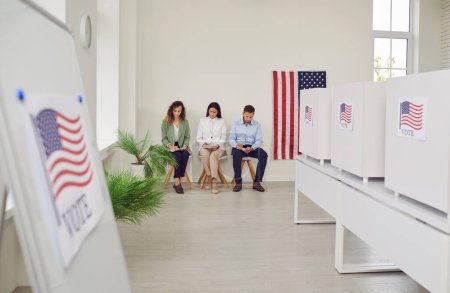 Polling station with a row of white voting booths decorated with American flag at vote center. People american voters sitting at polling station. Democracy and election day concept.