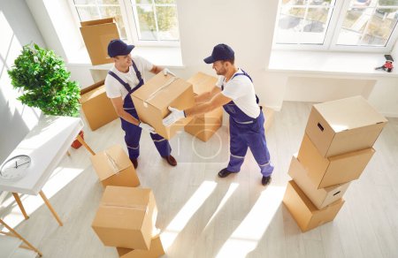 Photo for Top view of a team of young cheerful professional workers of moving service in overalls carrying and delivering cardboard boxes in new apartment. Move, moving day and relocation concept. - Royalty Free Image