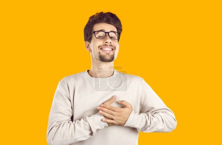Photo for Good-natured young guy with glasses has a friendly expression on his face, holds his hands on his heart, expresses his feelings, shows love and great sympathy. Isolated on a yellow background. - Royalty Free Image