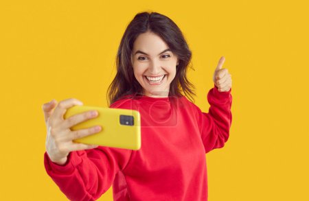 Téléchargez les photos : Joyeux rire jeune blogueuse brune en sweat-shirt rouge prend selfie sur smartphone montrant pouce vers le haut sur fond jaune. Appel vidéo, tournage vlog, pouce vers le haut geste aux abonnés. - en image libre de droit