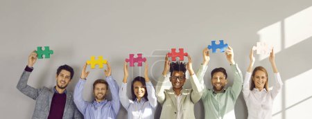Business-Team mit Puzzleteilen. Eine Gruppe fröhlicher multiethnischer Menschen steht an einer grauen Bürowand und hält bunte grüne, gelbe, rosa, rote, blaue und weiße Puzzleteile in die Höhe. Teamwork-Konzept