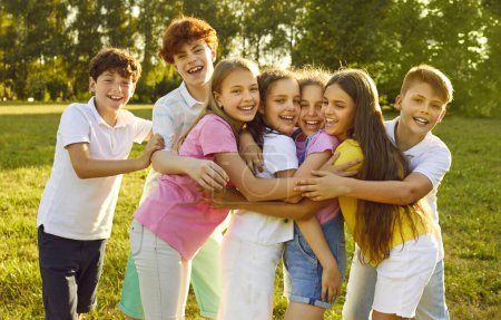 Téléchargez les photos : Portrait d'enfants heureux souriants amis debout sur l'herbe verte ensemble dans le parc à l'extérieur et regardant joyeusement la caméra étreignant et s'amusant. Camp d'été pour enfants et concept vacances. - en image libre de droit