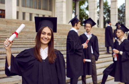 Photo for Portrait of young smiling joyful girl student in university graduate gown and diploma in hands. Happy graduated woman standing with classmates outdoor. Graduation celebration and education concept. - Royalty Free Image