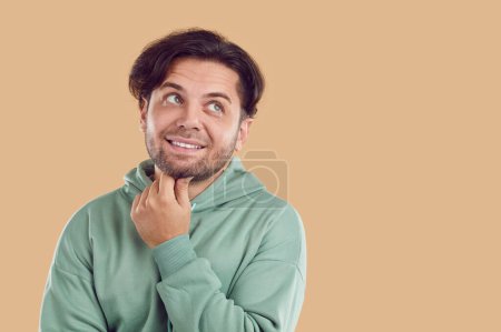 Thoughtful puzzled unshaved smiling brunet young handsome man in green sweatshirt looking up on copy space on beige background. He is touching his chin making decision or thinking about problem.