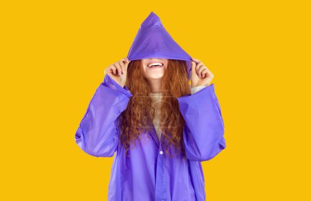Foto de Chica pelirroja rizada sonriente en impermeable púrpura escondida en la capucha en sus ojos sobre fondo amarillo. Ella está mirando hacia arriba a través de la capucha transparente en el cielo lluvioso. Traje de ropa para el clima lluvioso de otoño. - Imagen libre de derechos