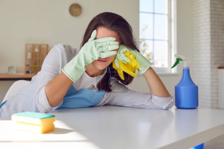 Portrait of a young tired woman housewife wearing rubber gloves with cleaning tools and rags, standing in kitchen and cover her face with hands. Housework, household or chores at home concept.
