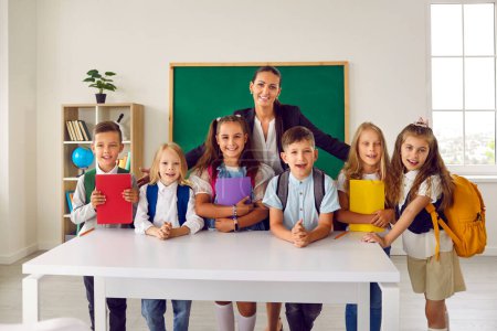 Group portrait of first graders with friendly female teacher in school classroom. Smiling children with backpacks and books stand in row with educator standing behind them. School concept.