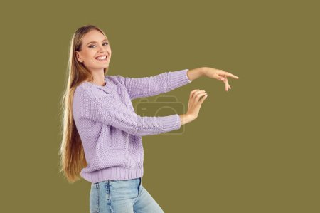 Photo for Smiling young woman, dressed in knitted sweater and blue jeans, dances, gracefully stretching her arms in front of her. Portrait of attractive blonde with long hair, side view on isolated background. - Royalty Free Image
