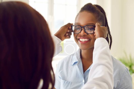 Téléchargez les photos : Examen des lunettes. Heureuse patiente afro-américaine essayant des lunettes comme recommandé par l'ophtalmologiste. Le docteur met des lunettes de vision noires sur la jeune femme. Concept d'ophtalmologie. - en image libre de droit