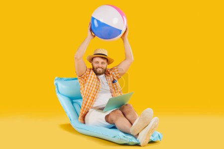 Photo for Funny happy man tourist in casual clothes sitting on inflatable mattress with beach ball booking tickets online via laptop on yellow background. Summer vacation, holiday trip or remote work concept - Royalty Free Image