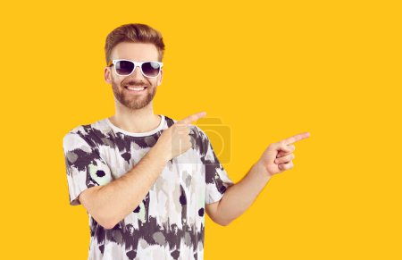 Téléchargez les photos : Homme joyeux dans les lunettes de soleil pointant vers le côté avec les deux index. Portrait d'un homme barbu confiant portant un t-shirt décontracté pointant avec les doigts pour copier l'espace sur fond jaune isolé - en image libre de droit
