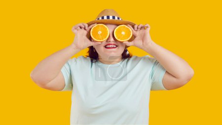 Foto de Retrato de verano de mujer gorda alegre divertida feliz usando sombrero de paja que cubre sus ojos con rodajas frescas de naranja aisladas en el fondo amarillo del estudio. Nutrición, dieta saludable y concepto de vacaciones. - Imagen libre de derechos