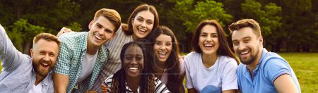 Photo for Happy adult friends enjoying summer and having fun together. Banner group portrait of cheerful joyful smiling young diverse multiracial people sitting on ground in park, with green trees in background - Royalty Free Image