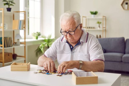 Portrait of a senior elderly sad gray-haired man collecting wooden jigsaw puzzles game at home sitting at the table. Memory training for dementia prevention and retirement leisure concept.