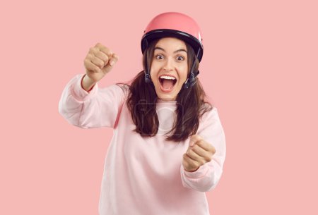 Photo for Crazy smiling amazed woman with opened mouth in helmet imagines she is driving and holding steering wheel on pink background. She is wearing pink sweatshirt. Human emotions, energy, drive concept. - Royalty Free Image