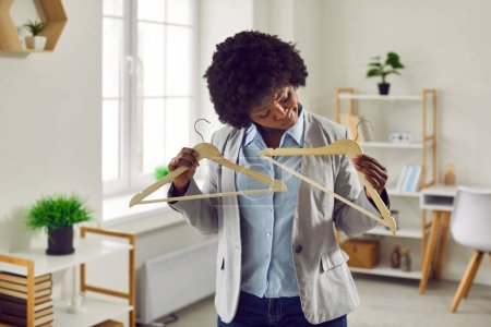 Photo for Doubting young woman holding two empty wooden hangers. Beautiful African American woman expressing nothing to wear from clothes or difficulty in deciding to change clothes - Royalty Free Image