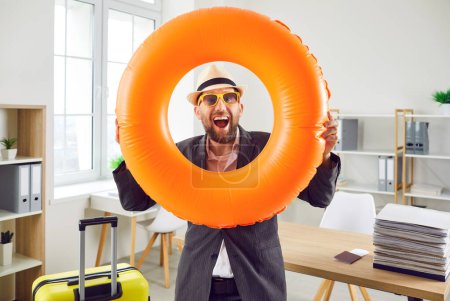 Photo for Joyful happy man holding yellow suitcase and swim ring while standing in office. Portrait of excited man wearing suit, summer hat and sunglasses looking through inflatable ring going on vacation - Royalty Free Image