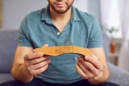 Photo for Cropped photo of a young smiling man wearing casual clothes sitting on sofa and holding orthopedic shoe insole in his hands at home. Feet comfort and health care concept. Selective focus. - Royalty Free Image