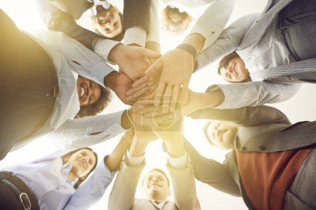 Eine Gruppe unterschiedlicher Geschäftsleute wird aus einem niedrigen Winkel gezeigt, wie sie im Kreis stehen und ihre Hände übereinander stapeln. Symbol für Teamfähigkeit, Teamarbeit und Zusammenarbeit im geschäftlichen Umfeld