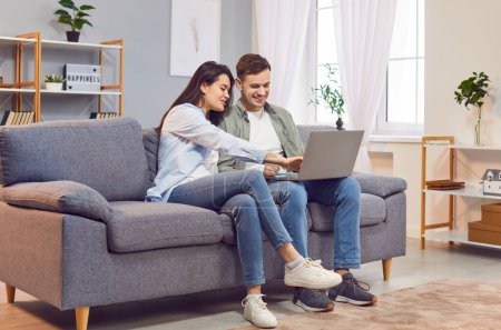 Photo for Young married couple on the couch in the living room, using a laptop together for online entertainment. They are happy and engaged, enjoying leisure time at home, talking and surfing the internet. - Royalty Free Image
