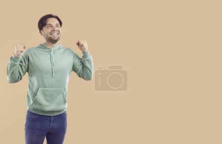 Téléchargez les photos : Jeune homme heureux souriant amusant en vêtements décontractés faire geste gagnant serrant les poings célébrant le succès isolé sur fond beige studio avec espace de copie. Concept d'émotions humaines. - en image libre de droit