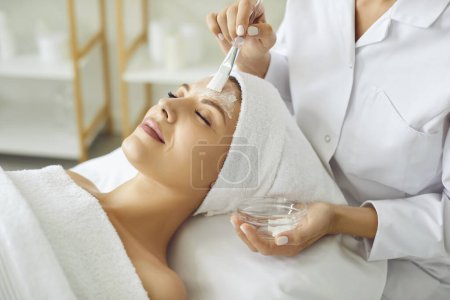 Téléchargez les photos : Cosmetologue appliquer un masque hydratant avec pinceau à une jeune femme souriante détendue dans le salon de spa. Gros plan portrait d'une cliente recevant une procédure de beauté de l'esthéticienne en clinique de beauté - en image libre de droit