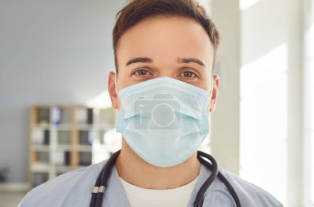Photo for Close-up portrait of young man doctor wearing medical mask with stethoscope looking at camera ready to help patients standing in clinic or hospital. Medicine and health care concept. - Royalty Free Image