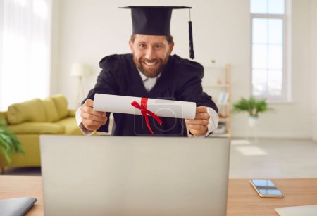 Happy graduate man showing his diploma via laptop having online video call. Male student sitting at the desk at home in gown in a virtual ceremony. Online degree and university education concept.
