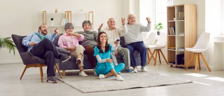 Photo for Group of senior people watching TV in retirement home together with young nurse. Active cheerful senior people sit on sofa and shout with joy when nurse switches channels with remote control. - Royalty Free Image