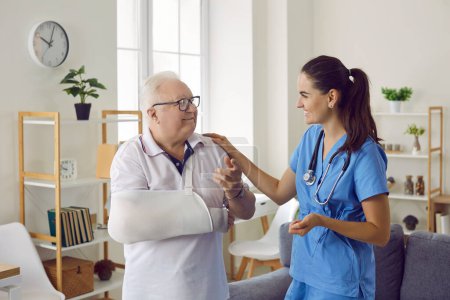 Doctor supporting her senior patient with an arm injury. Friendly young woman in blue uniform scrubs talking to a happy old man with a broken arm. Medical help, injury treatment concept