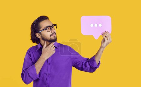 Studio shot of thoughtful Indian man with message bubble. Handsome ethnic guy in purple shirt and glasses standing isolated on yellow background, looking at chat message with three dots and thinking